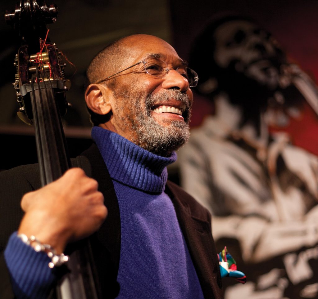 Cette image montre un homme souriant, probablement un musicien, tenant une contrebasse. Il porte un pull à col roulé bleu et une veste noire, avec une pochette colorée dans sa poche de veste. Son expression joyeuse et confiante, ainsi que le cadre artistique en arrière-plan, suggèrent qu'il pourrait partager un témoignage sur son expérience en tant que contrebassiste. Il pourrait évoquer l'importance d'un bon archet en carbone pour obtenir un son riche et précis, et comment cela a influencé sa carrière musicale.