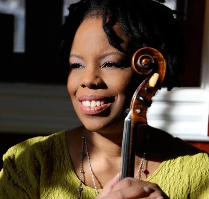 Cette image montre une femme souriante, tenant un violon. Elle est éclairée par une lumière naturelle qui met en valeur son visage serein et ses cheveux noirs. Son témoignage pourrait porter sur l'importance de la musique dans sa vie et la précision nécessaire pour jouer avec un archet en carbone, qui offre une résonance et une sensibilité exceptionnelles.