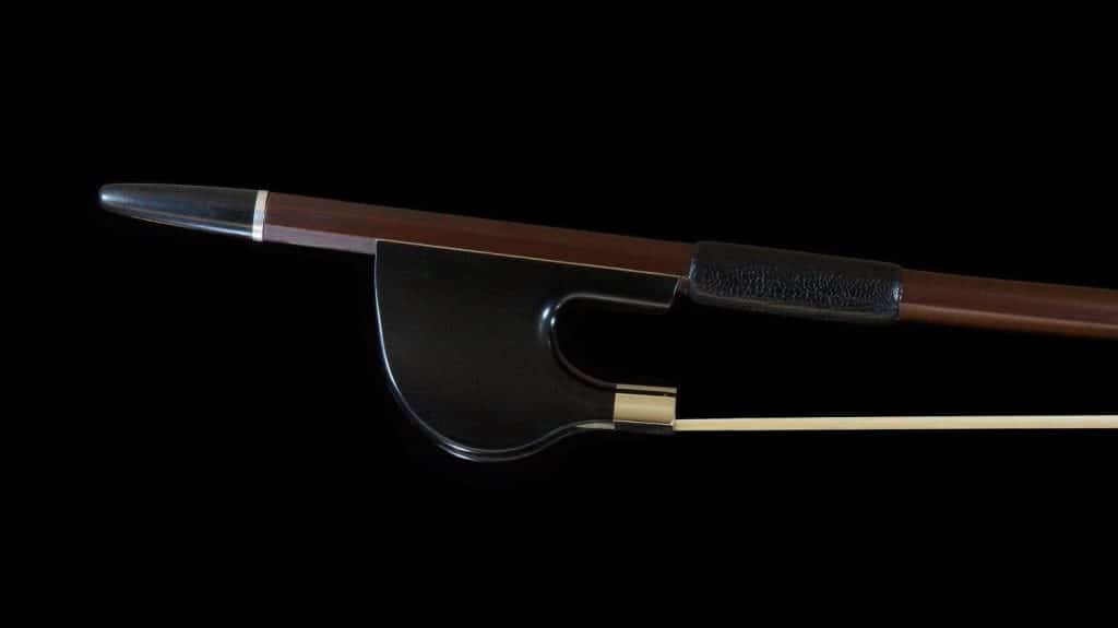Cette image montre un archet en carbone de profil, posé sur un fond noir, mettant en valeur son design élégant et moderne. La baguette est brune avec un grip noir, et la hausse en carbone a une finition lisse et brillante. L'archet est finement fabriqué, combinant tradition et technologie moderne pour offrir une performance optimale aux musiciens exigeants.